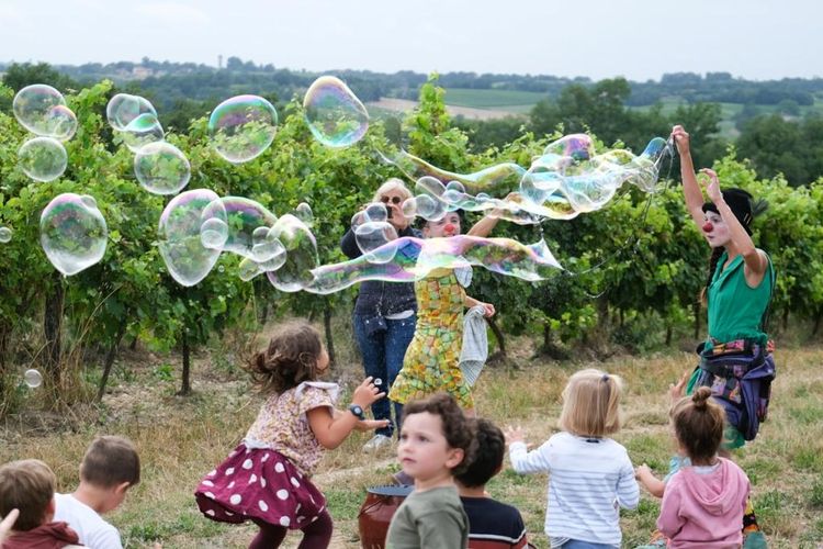 Une femme avec un nez de clown fait d'énormes bulles de savon près d'une vigne, avec des enfants qui essaient de les attraper